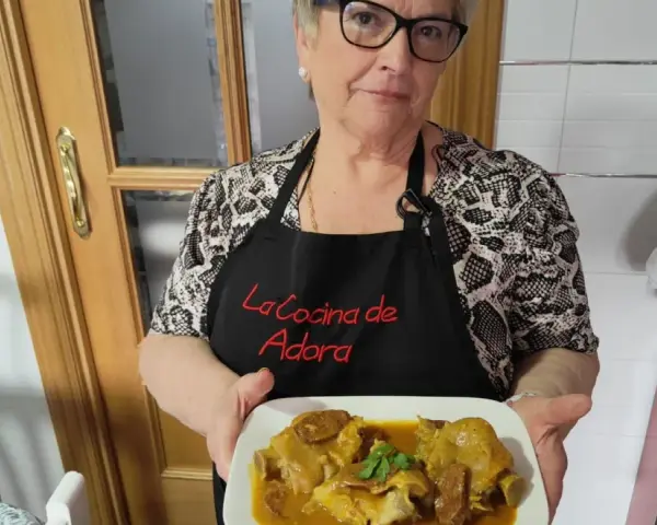 Una mujer sonriente muestra un plato de manitas de cerdo en salsa. Lleva un delantal negro con el texto "La Cocina de Adora".