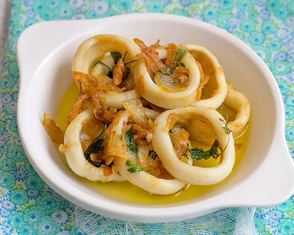 Calamares encebollados en un plato blanco, con cebolla caramelizada y perejil, sobre un mantel azul y verde.