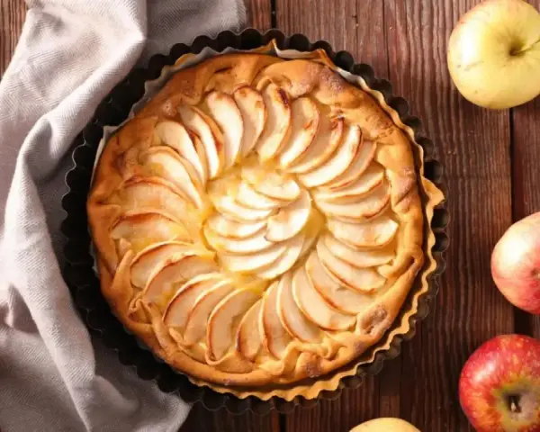 Tarta de manzana dorada, decorada con láminas de fruta, rodeada de manzanas frescas y espigas de trigo sobre una mesa de madera.