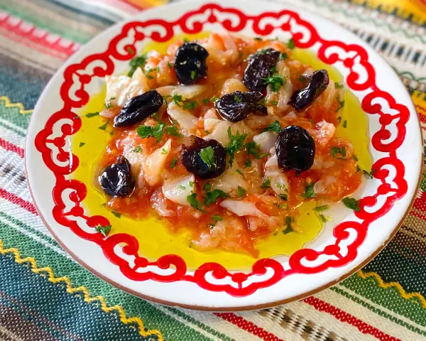 Esqueixada de bacalao con aceitunas negras y perejil, servida en un plato blanco con borde rojo sobre un mantel colorido.