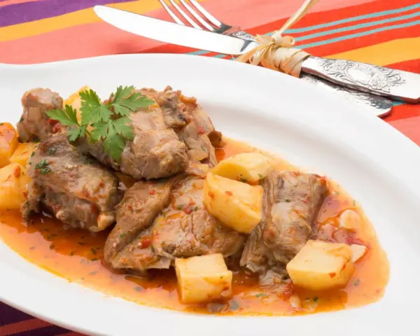 Sabroso cordero al chilindrón con patatas, servido en plato blanco con cubiertos atados con hilo.