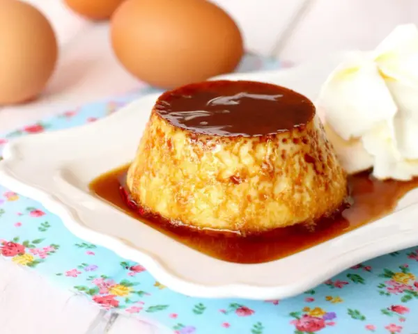 Delicioso flan de huevo casero con caramelo líquido y una porción de nata montada. Huevos frescos al fondo.
