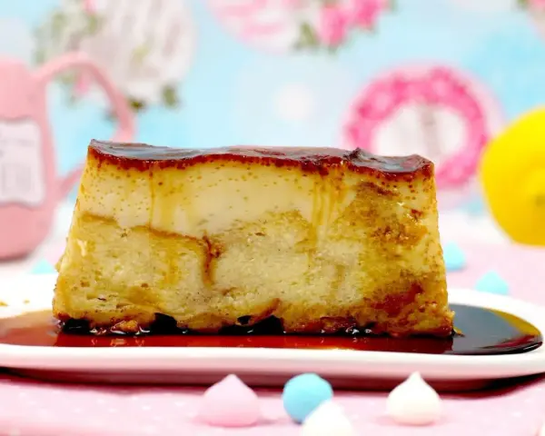 Un trozo de pan de calatrava, con su caramelo dorado, reposa en un plato blanco. Al fondo, una taza rosa y un limón.