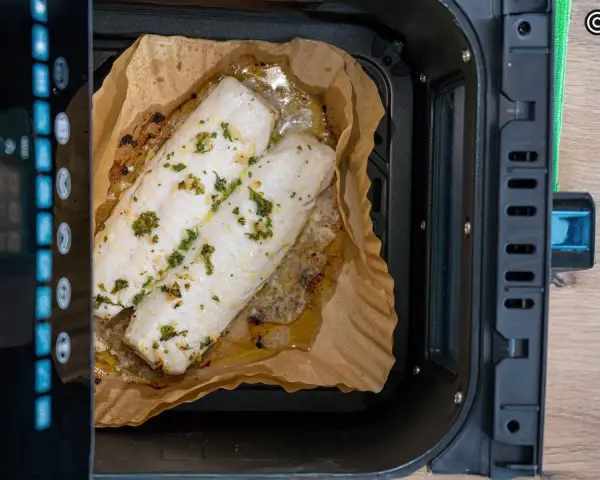 Dos filetes de merluza con hierbas cocinándose en papel de horno dentro de una freidora de aire.