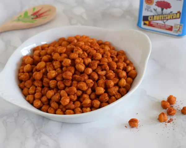 Un tazón blanco lleno de garbanzos fritos crujientes y especiados. Una lata de clavo y una cuchara de madera decorada están al fondo.