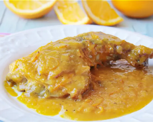 Delicioso pollo a la naranja, bañado en una salsa dorada y jugosa, con gajos de naranja frescos al fondo.