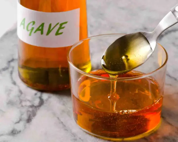 Una cuchara vierte sirope de agave dorado en un vaso lleno de este dulce líquido. Una botella con etiqueta "AGAVE" está al fondo.