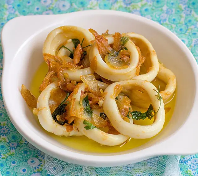 Calamares encebollados en un plato blanco, con cebolla caramelizada y perejil, sobre un mantel azul y verde.