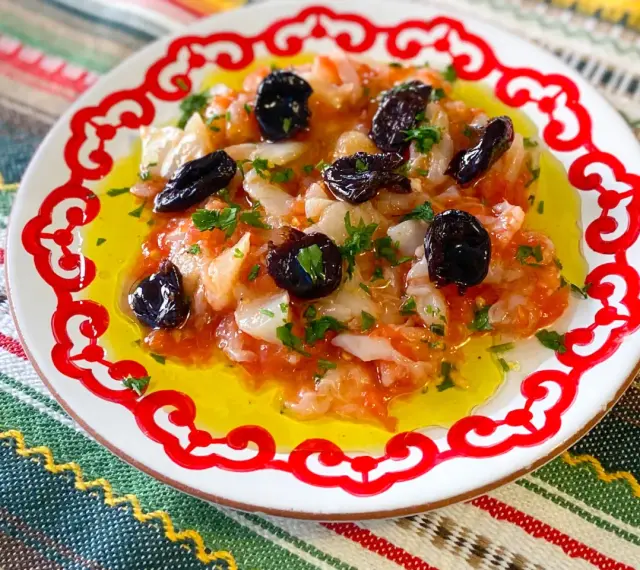 Esqueixada de bacalao con aceitunas negras y perejil, servida en un plato blanco con borde rojo sobre un mantel colorido.