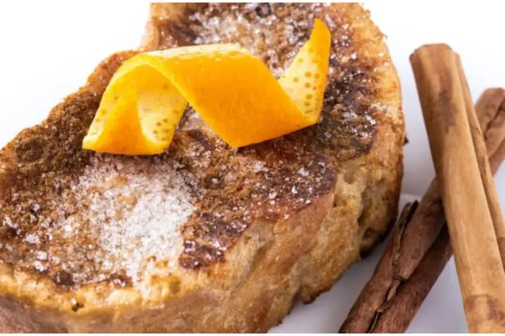 Torrijas al horno espolvoreadas con azúcar y canela, decoradas con piel de naranja y acompañadas de ramas de canela.