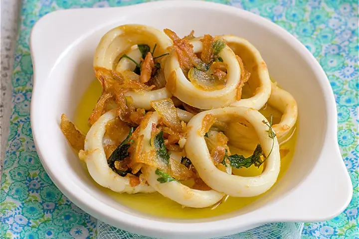 Calamares encebollados en un plato blanco, con cebolla caramelizada y perejil, sobre un mantel azul y verde.