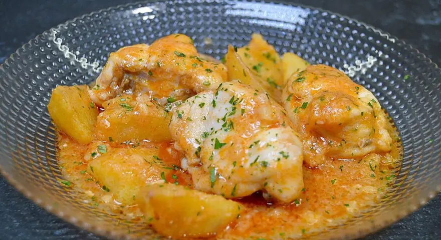 Rape a la marinera con patatas, servido en un cuenco de cristal con salsa naranja y perejil picado.