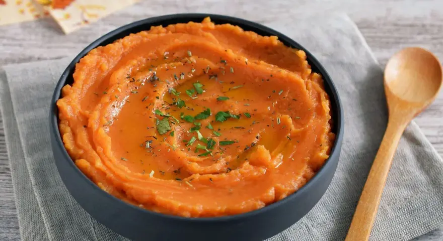 Un tazón de cremoso puré de boniato, adornado con aceite y hierbas, junto a una cuchara de madera.