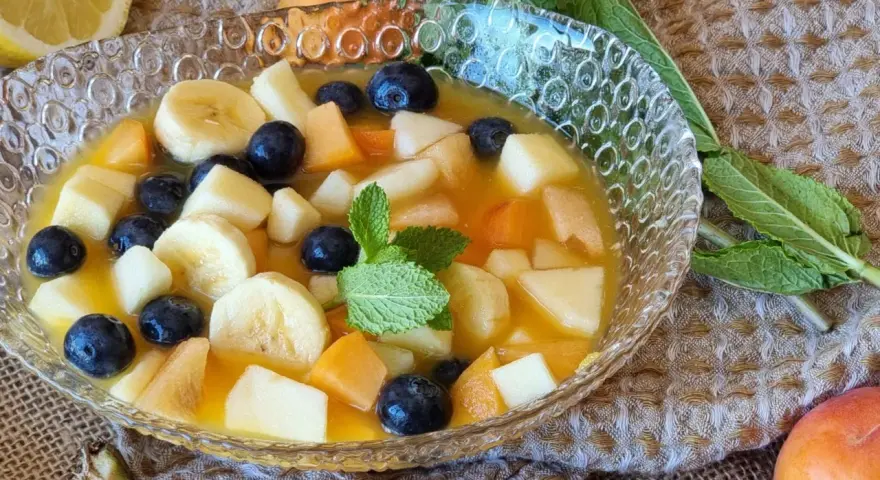 Deliciosa macedonia de frutas con plátano, arándanos y melocotón en un cuenco de cristal, decorada con menta.