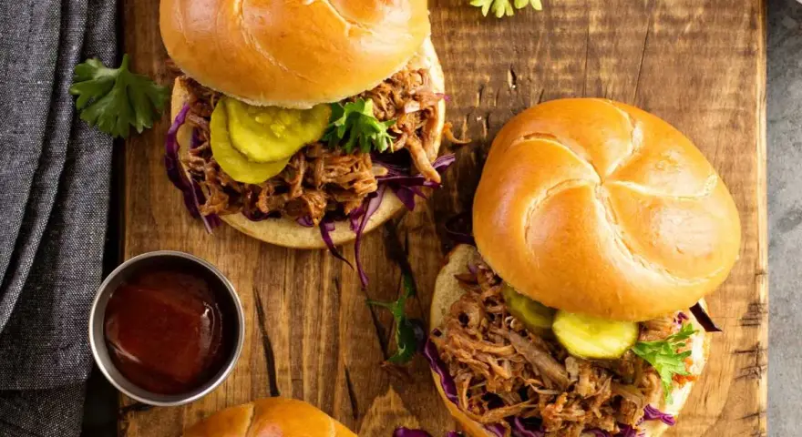 Dos sándwiches de pulled pork jugoso, con repollo morado, pepinillos y salsa barbacoa en un plato de madera.