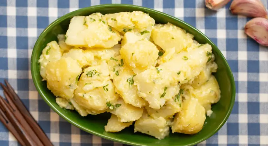 Un tazón verde lleno de patatas alioli cremosas, adornadas con perejil fresco. A su lado, ajos y tenedores de madera sobre un mantel de cuadros azules.