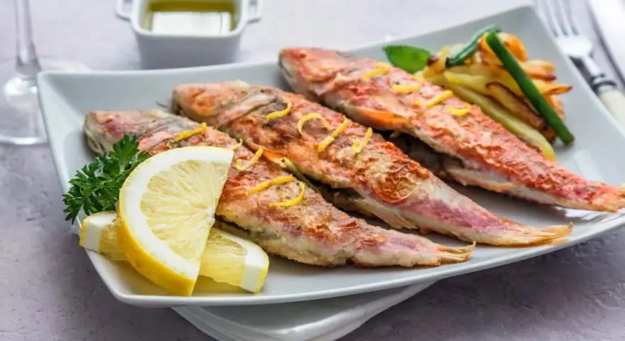 Cuatro salmonetes al horno, dorados y jugosos, servidos con rodajas de limón, perejil y patatas fritas.