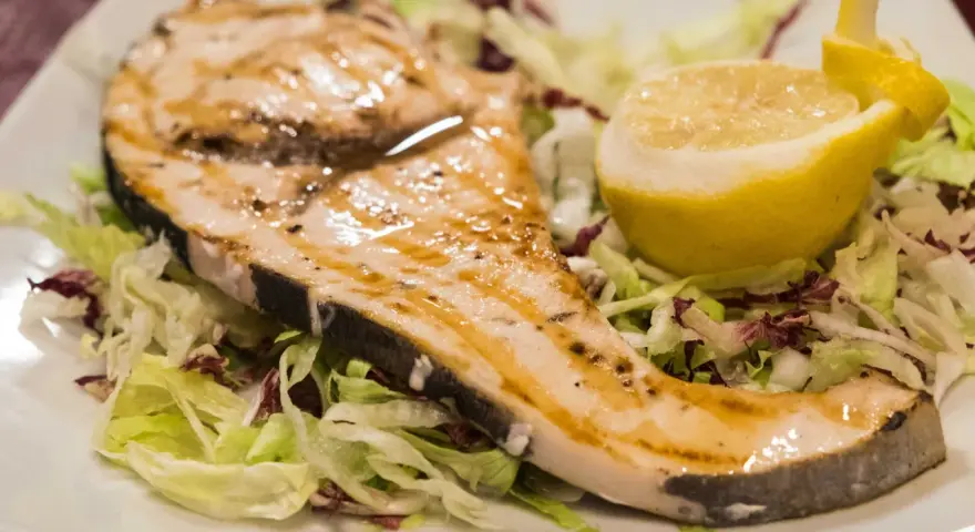 Pez espada a la plancha con ajo y perejil, servido sobre una cama de lechugas y acompañado de un gajo de limón.