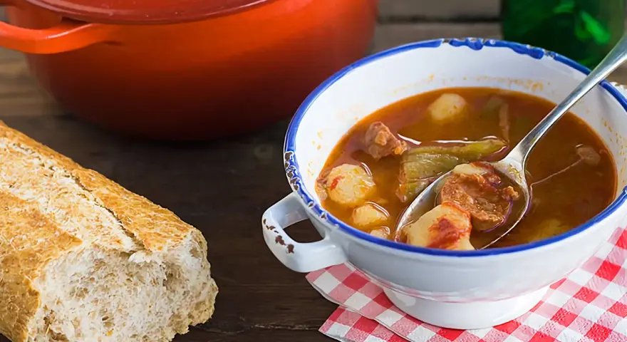 Un cuenco de patatas a la riojana humeante, con chorizo y pimientos, junto a una hogaza de pan rústico y una olla roja.