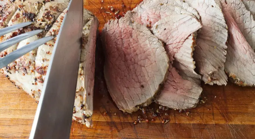 Un cuchillo corta un jugoso roast beef en rodajas sobre una tabla de madera.