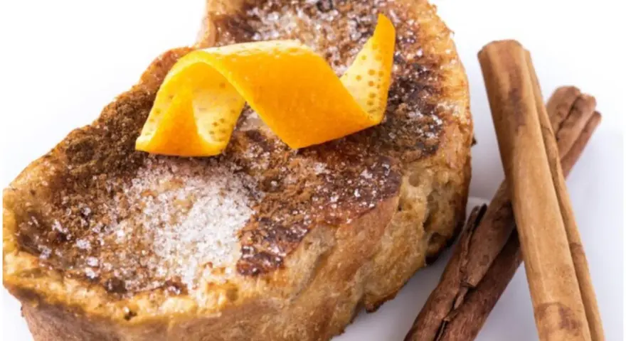 Torrijas al horno espolvoreadas con azúcar y canela, decoradas con piel de naranja y acompañadas de ramas de canela.