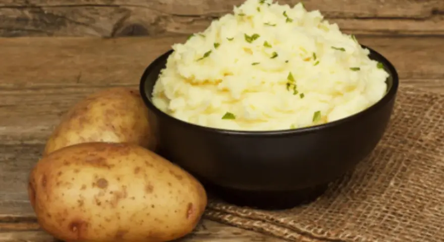 Un tazón de cremoso puré de patatas, adornado con perejil picado, junto a patatas enteras sobre una mesa de madera rústica.