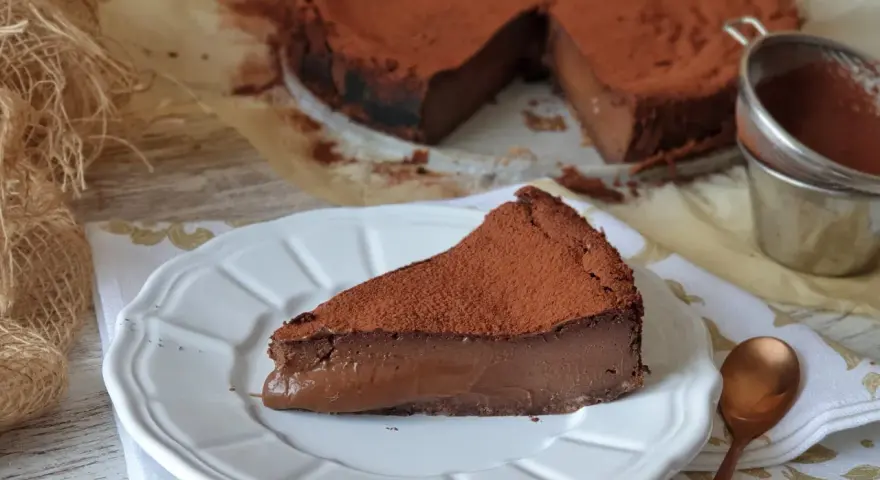 Una porción de tarta de chocolate, cubierta de cacao en polvo, reposa en un plato blanco. El resto de la tarta, también espolvoreada, se ve al fondo.