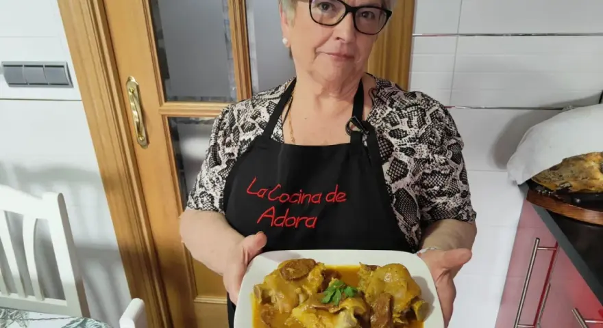 Una mujer sonriente muestra un plato de manitas de cerdo en salsa. Lleva un delantal negro con el texto "La Cocina de Adora".