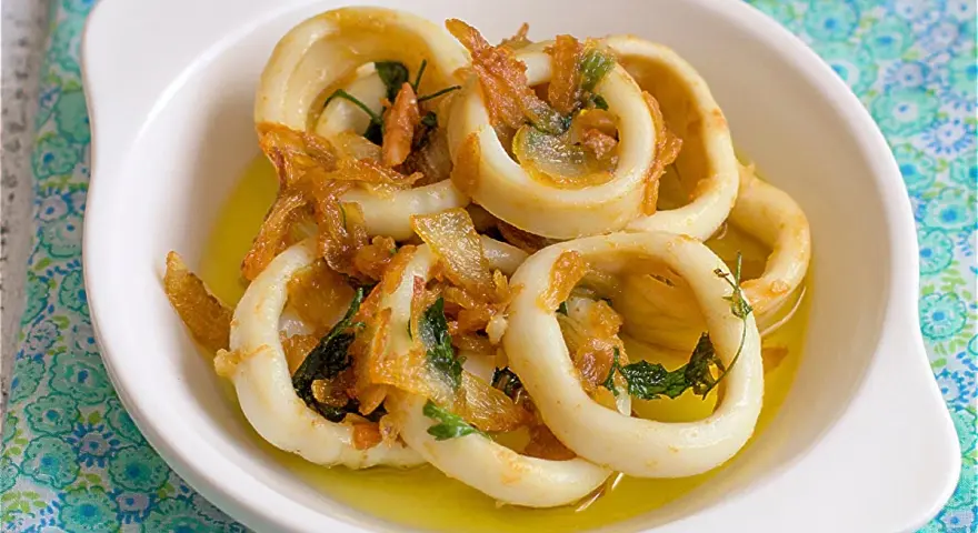 Calamares encebollados en un plato blanco, con cebolla caramelizada y perejil, sobre un mantel azul y verde.