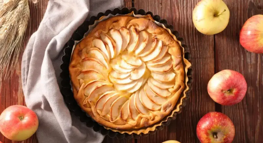 Tarta de manzana dorada, decorada con láminas de fruta, rodeada de manzanas frescas y espigas de trigo sobre una mesa de madera.