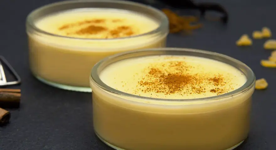 Dos cuencos de natillas caseras, espolvoreadas con canela, junto a palitos de canela y vainilla.