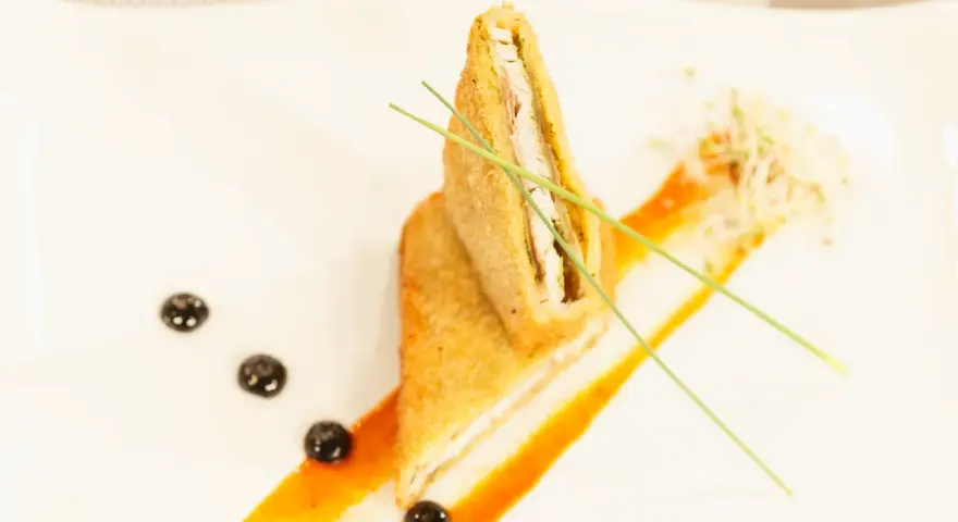 Un crujiente san jacobo, cortado por la mitad, reposa sobre una salsa naranja. Brotes y puntos oscuros decoran el plato.