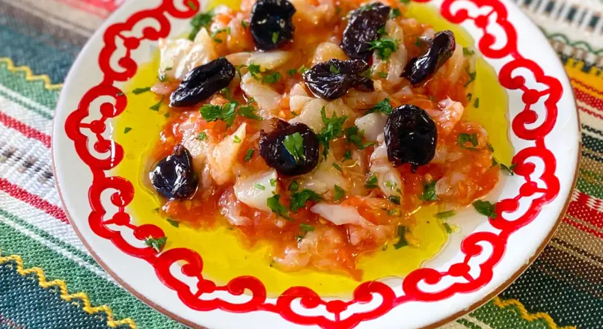 Esqueixada de bacalao con aceitunas negras y perejil, servida en un plato blanco con borde rojo sobre un mantel colorido.