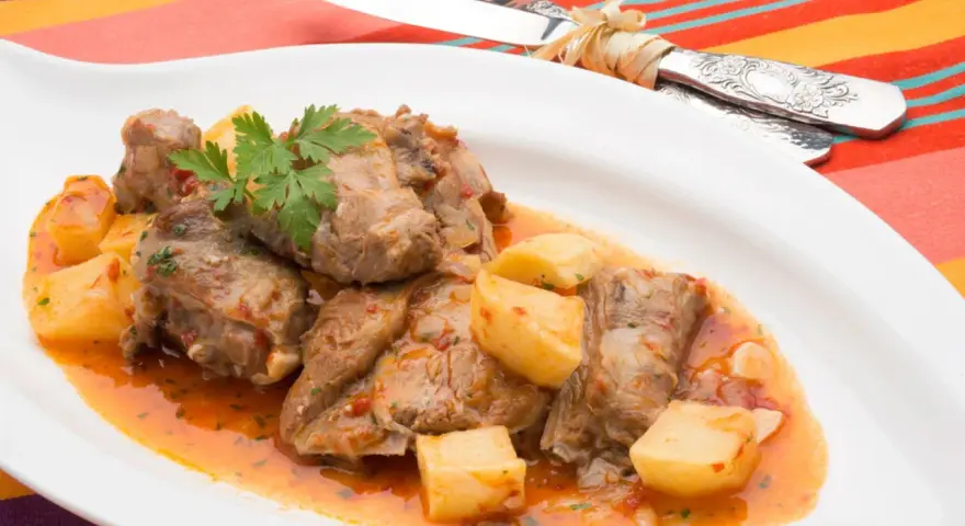 Sabroso cordero al chilindrón con patatas, servido en plato blanco con cubiertos atados con hilo.