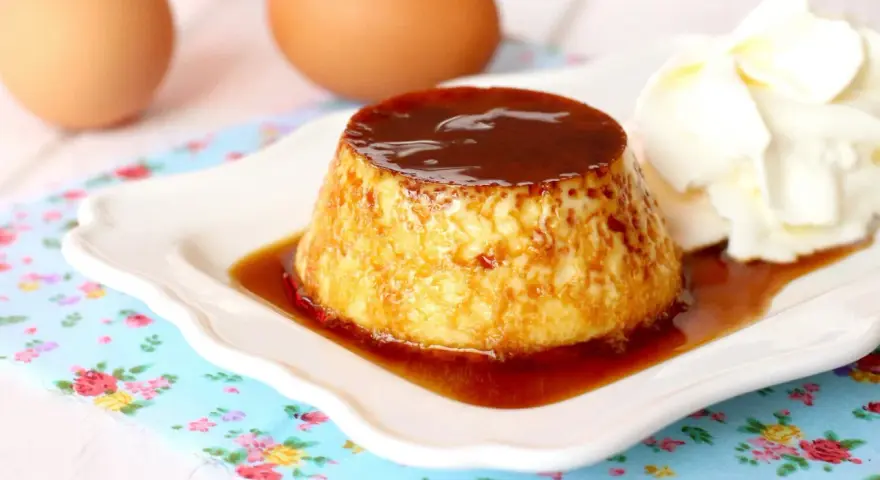 Delicioso flan de huevo casero con caramelo líquido y una porción de nata montada. Huevos frescos al fondo.