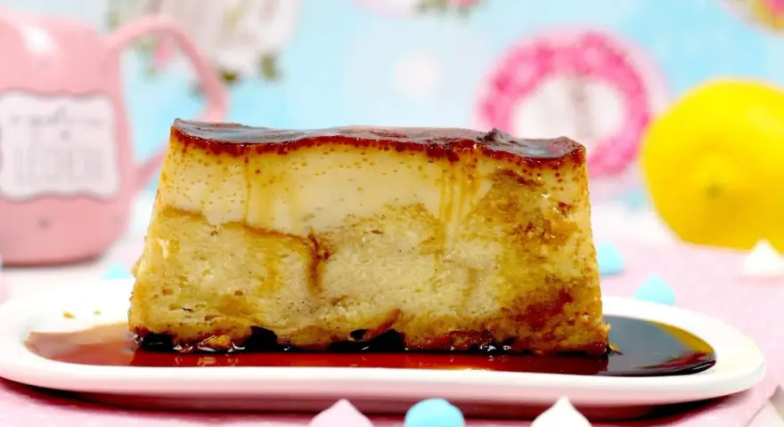 Un trozo de pan de calatrava, con su caramelo dorado, reposa en un plato blanco. Al fondo, una taza rosa y un limón.