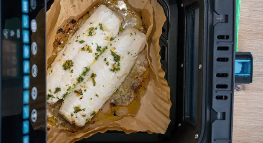 Dos filetes de merluza con hierbas cocinándose en papel de horno dentro de una freidora de aire.
