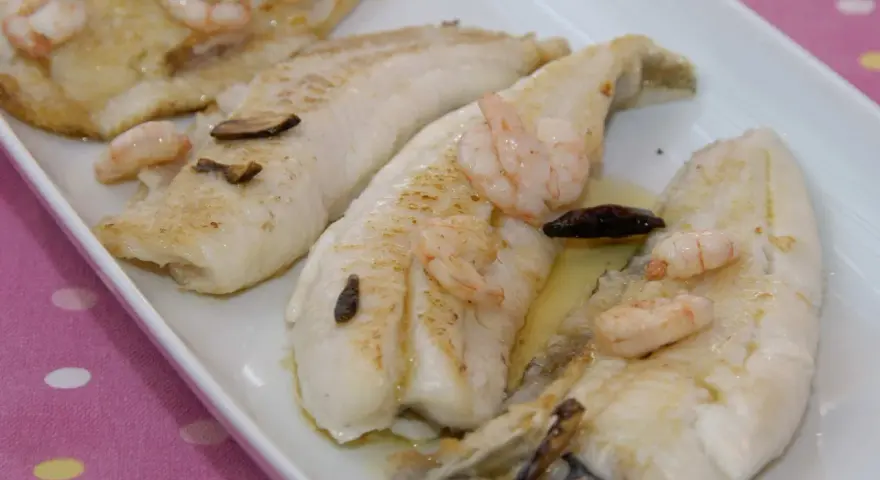 Filetes de gallo a la plancha con gambas y ajo laminado, servidos en una bandeja blanca.