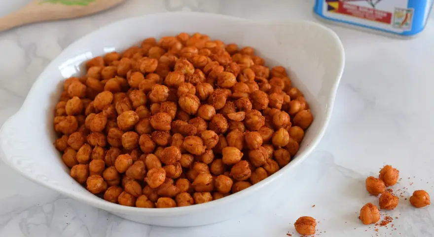 Un tazón blanco lleno de garbanzos fritos crujientes y especiados. Una lata de clavo y una cuchara de madera decorada están al fondo.