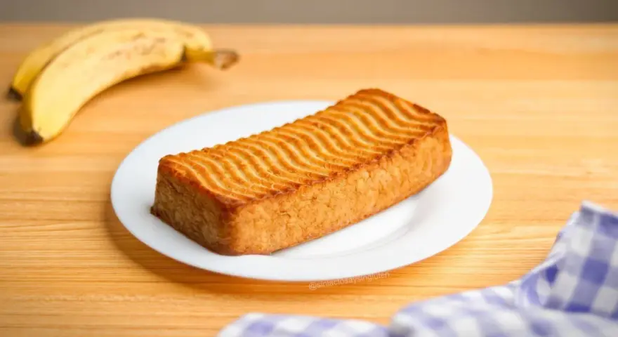 Un delicioso bizcocho de plátano con un patrón ondulado dorado en la parte superior, acompañado de plátanos maduros.