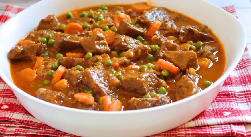 Ternera a la jardinera en un cuenco blanco, con trozos de carne tierna, zanahorias y guisantes en una salsa sabrosa.