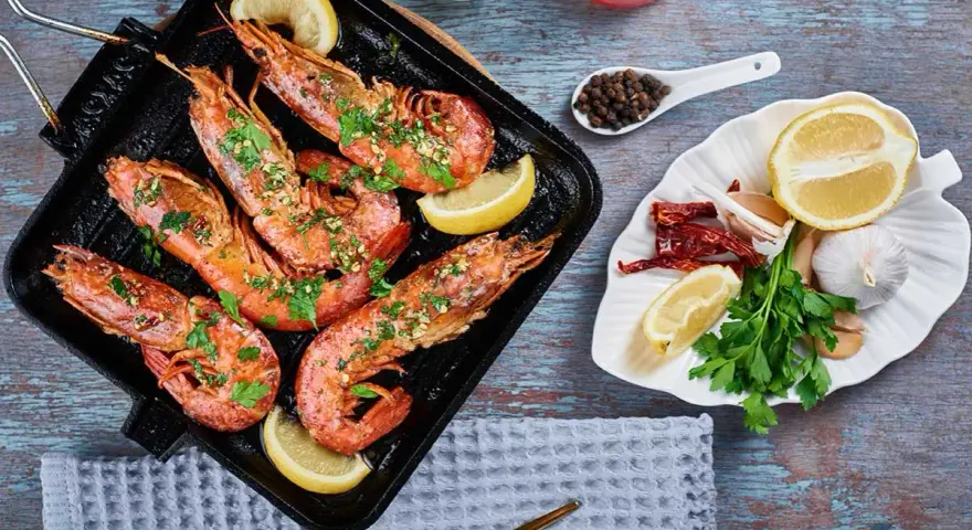 Deliciosos langostinos a la plancha, servidos con rodajas de limón y perejil fresco. Acompañados de ajo, guindillas y granos de pimienta.