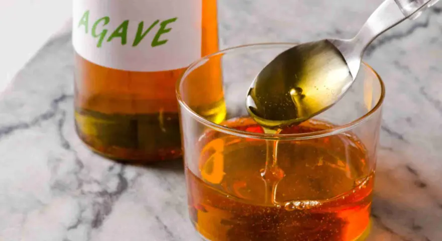 Una cuchara vierte sirope de agave dorado en un vaso lleno de este dulce líquido. Una botella con etiqueta "AGAVE" está al fondo.