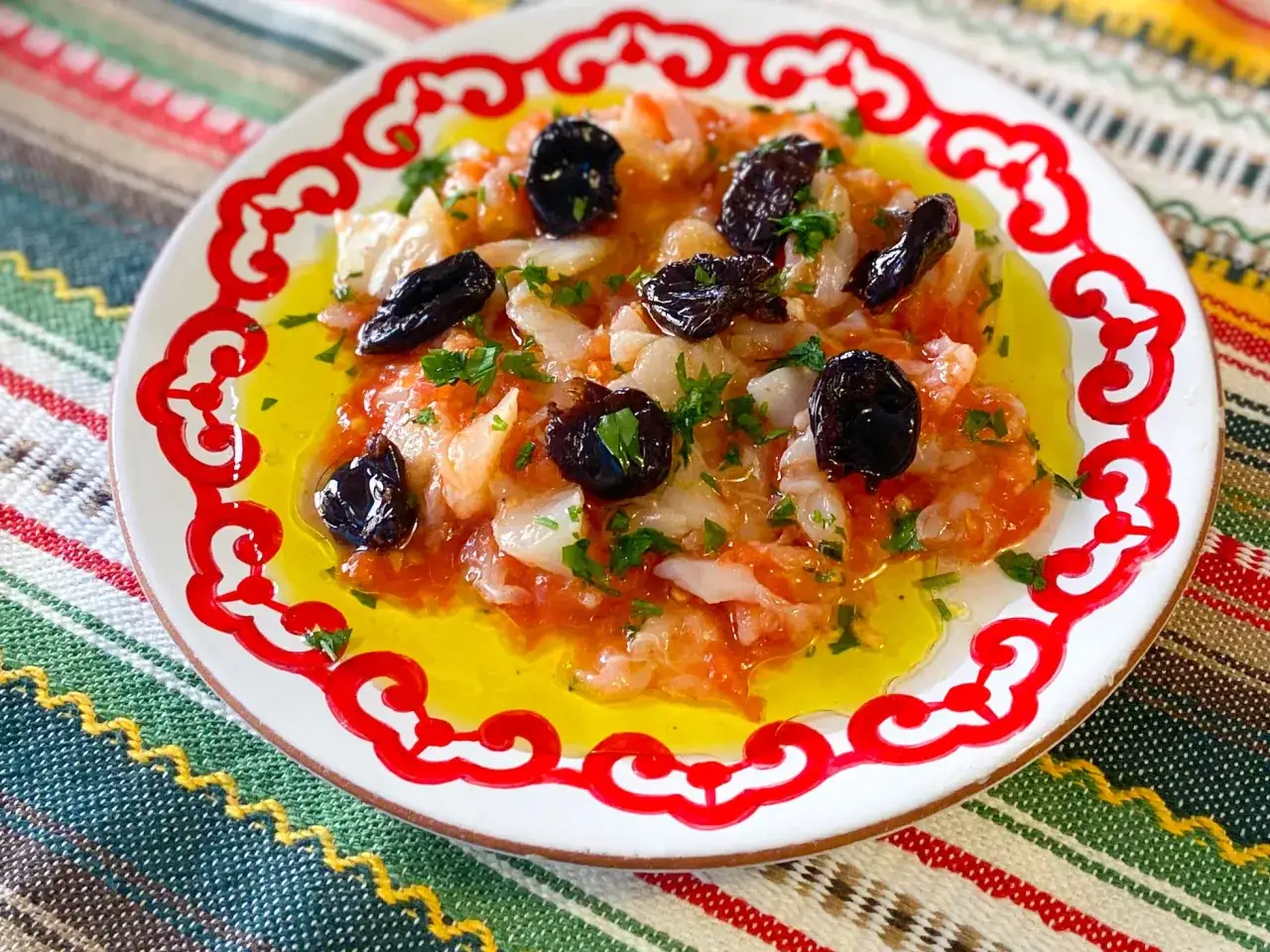 Esqueixada de bacalao con aceitunas negras y perejil, servida en un plato blanco con borde rojo sobre un mantel colorido.