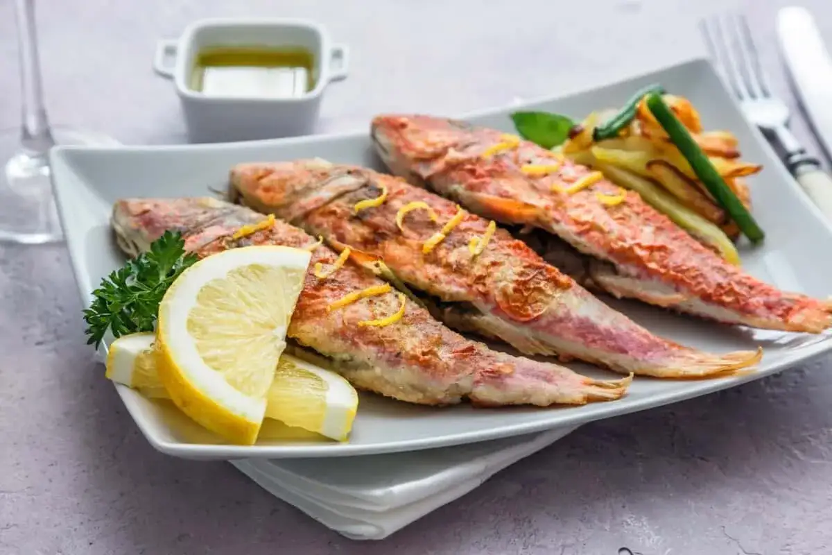 Cuatro salmonetes al horno, dorados y crujientes, servidos con rodajas de lim&oacute;n, perejil y patatas fritas.