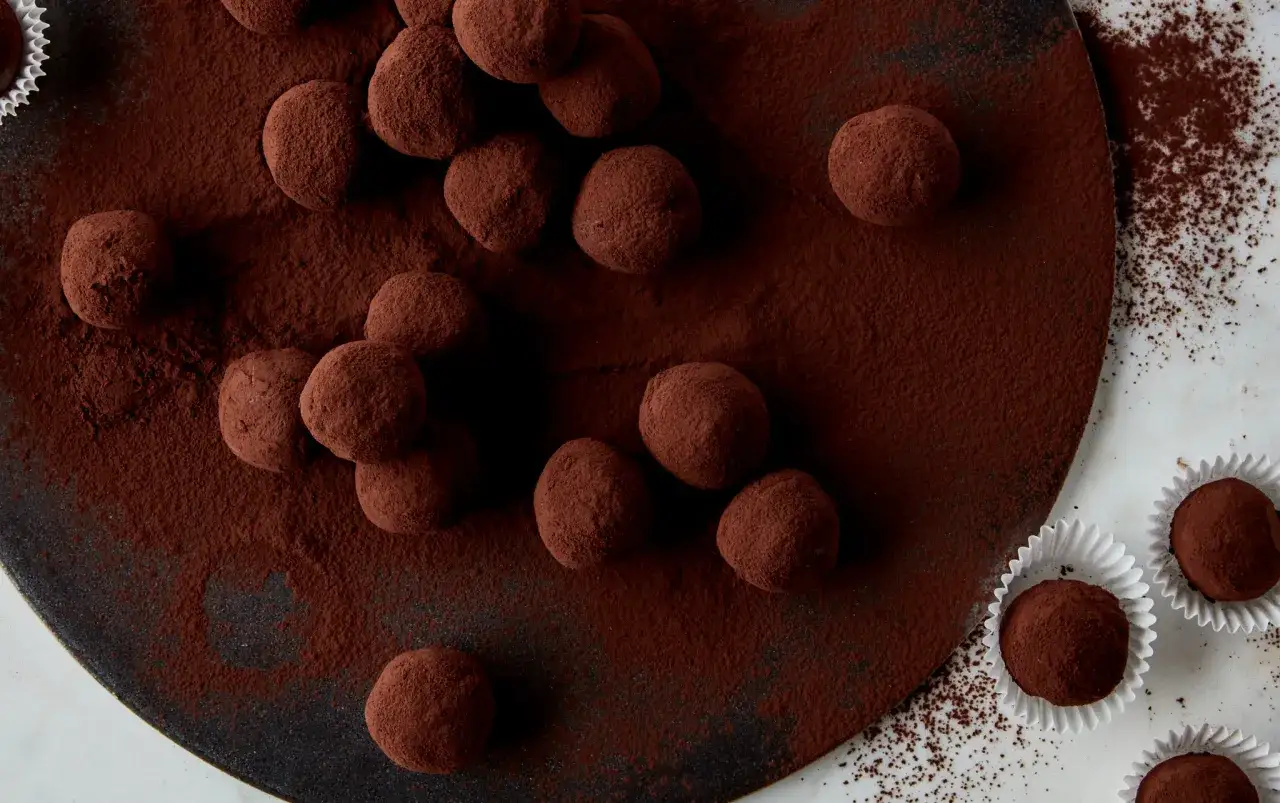 Mont&oacute;n de trufas de chocolate espolvoreadas con cacao en polvo sobre una bandeja oscura. Algunas trufas est&aacute;n en capacillos blancos.