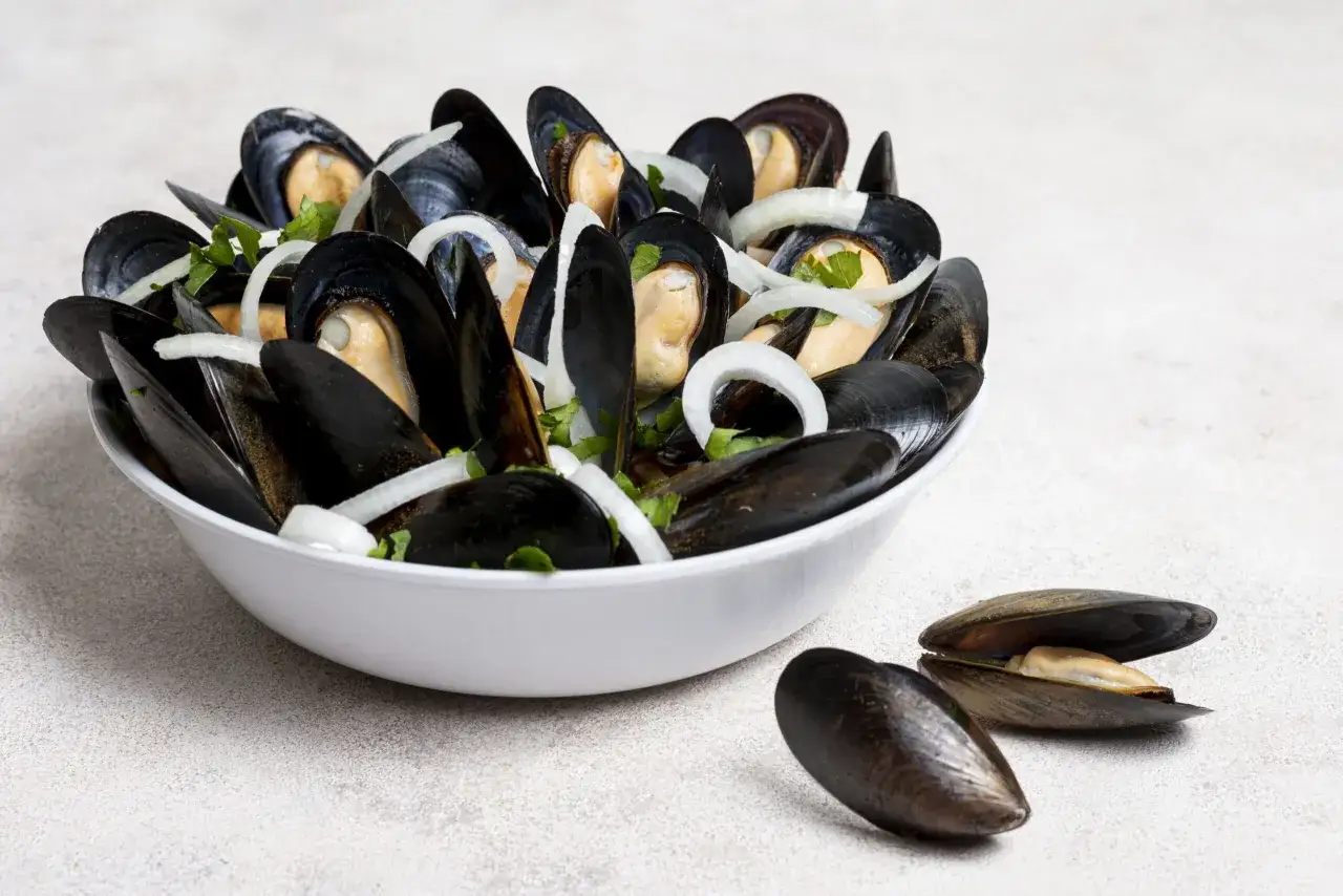 Un taz&oacute;n blanco lleno de mejillones al vapor, adornados con cebolla y perejil. Dos mejillones abiertos descansan al frente.