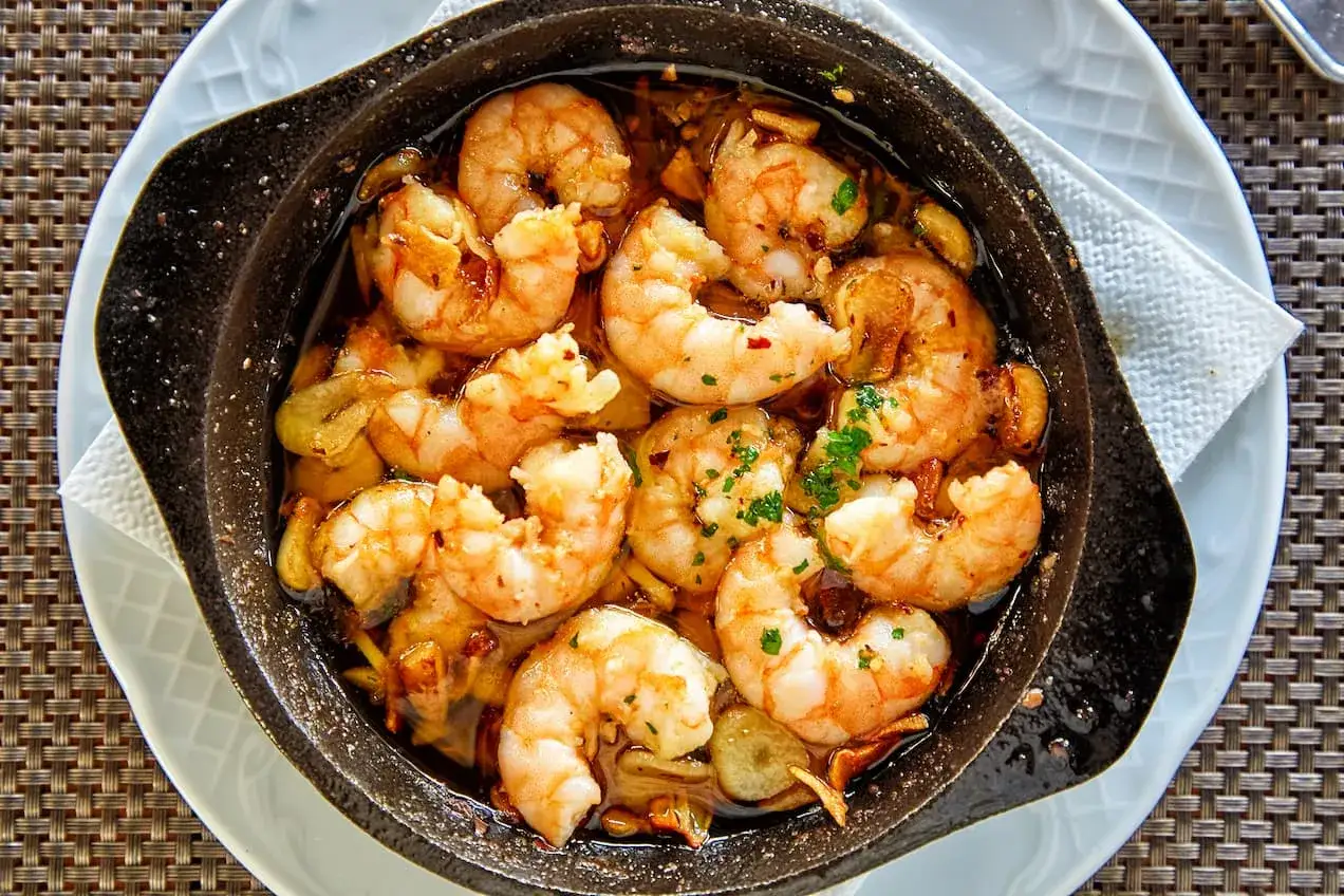 Gambones al ajillo burbujeantes en una cazuela de barro, con ajo laminado y perejil picado.