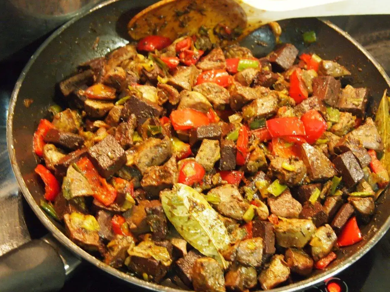 Frito mallorquín en sartén, con trozos de carne, pimientos rojos y verdes, y hojas de laurel.