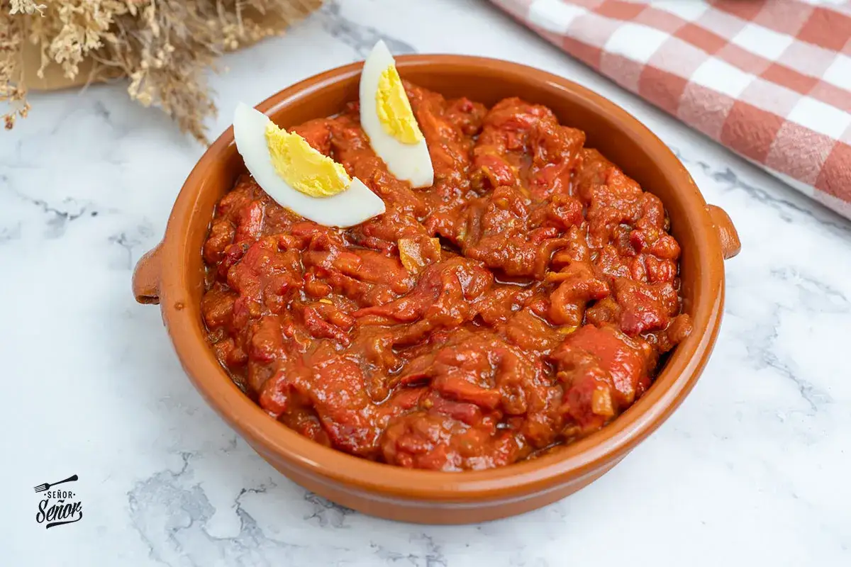 Un plato de asadillo manchego, con pimientos rojos guisados y trozos de huevo cocido, servido en una cazuela de barro.